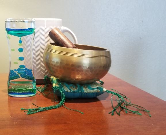 Image of brass singing bowl on small green pillow with tassles, a bubble timer, and a white mug in the background