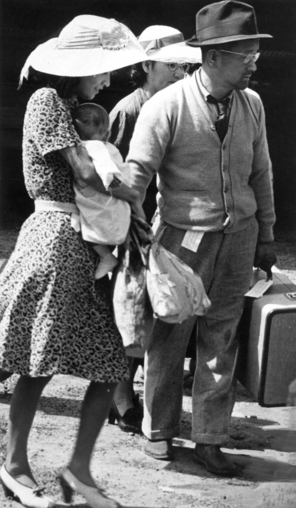  Japanese family on their way to the Granada Relocation Center, Camp Amache, Prowers County, southeastern Colorado, includes two women in dresses, heels and bonnets. One holds a baby, and a man in a sweater, tie, eye glasses and a hat, holds a suitcase. The man has a tag on his belt and another on the suitcase.