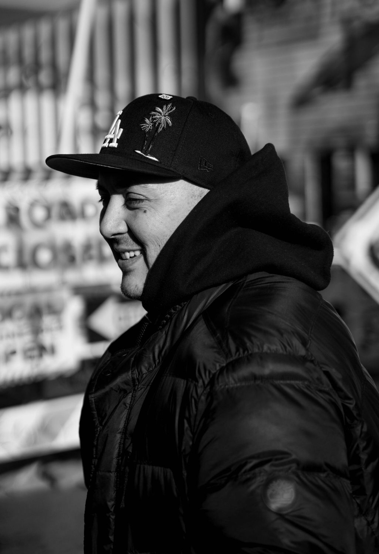 Black and white photograph of the presenting artist. He is centered in the frame, alone, smiling. The image is shot profile and he is wearing a baseball hat and jacket.