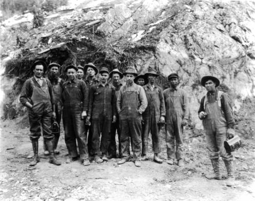 Miners, some possibly Native American or Asian, pose outdoors in Ouray County, Colorado. Some men hold lunch boxes.