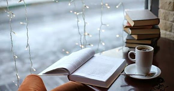A wooden table with an open book, a mug of coffee, and a stack of books sits in front of a window.