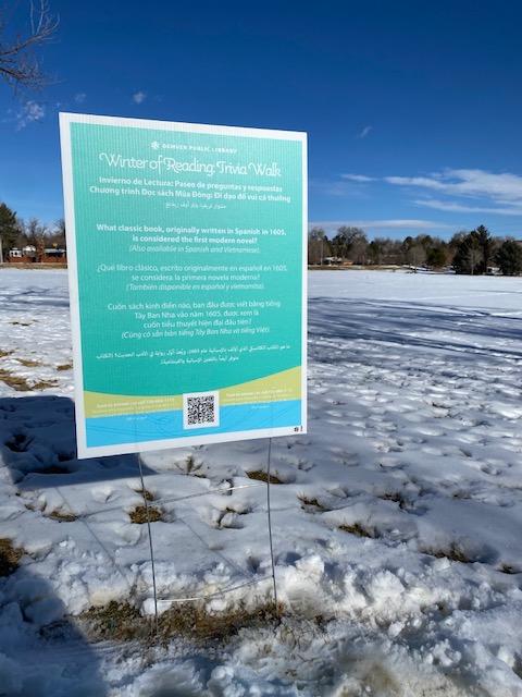 A large blue sign that reads "Winter of Reading Trivia Walk" staked into the ground of a snow-covered park.