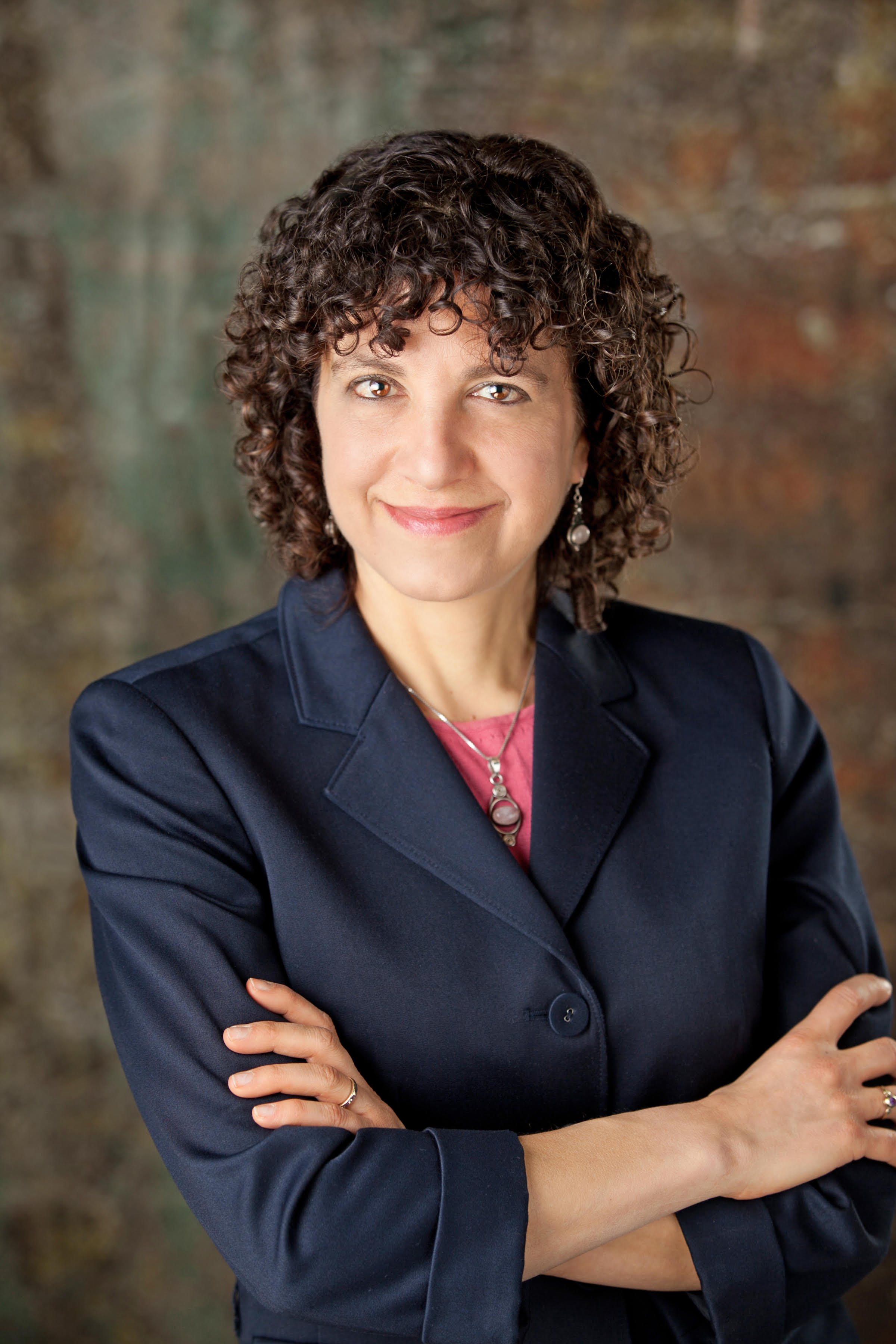 Becca Levy, a woman with curly brown hair, brown eyes, and light skin in a blue blazer and pink shirt with her arms crossed.