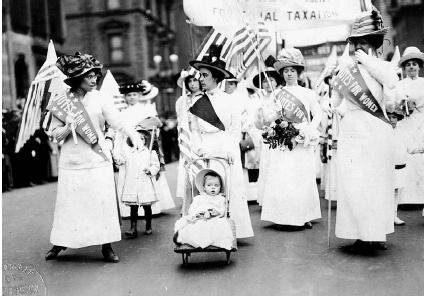 Women in white dresses marching in a parade, holding signs that say "Votes for Women"