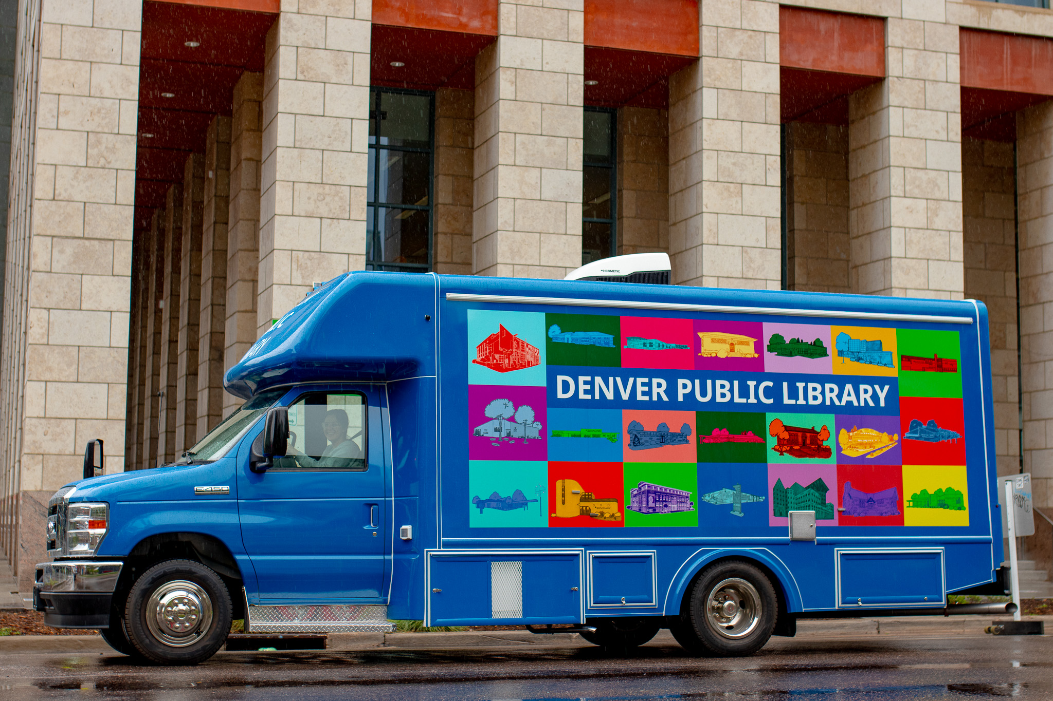 A bright blue Denver Public Library bookmobile parked in front of a large stone building on a rainy day. The side of the vehicle features a colorful grid of illustrated library locations and a large ‘Denver Public Library’ logo.