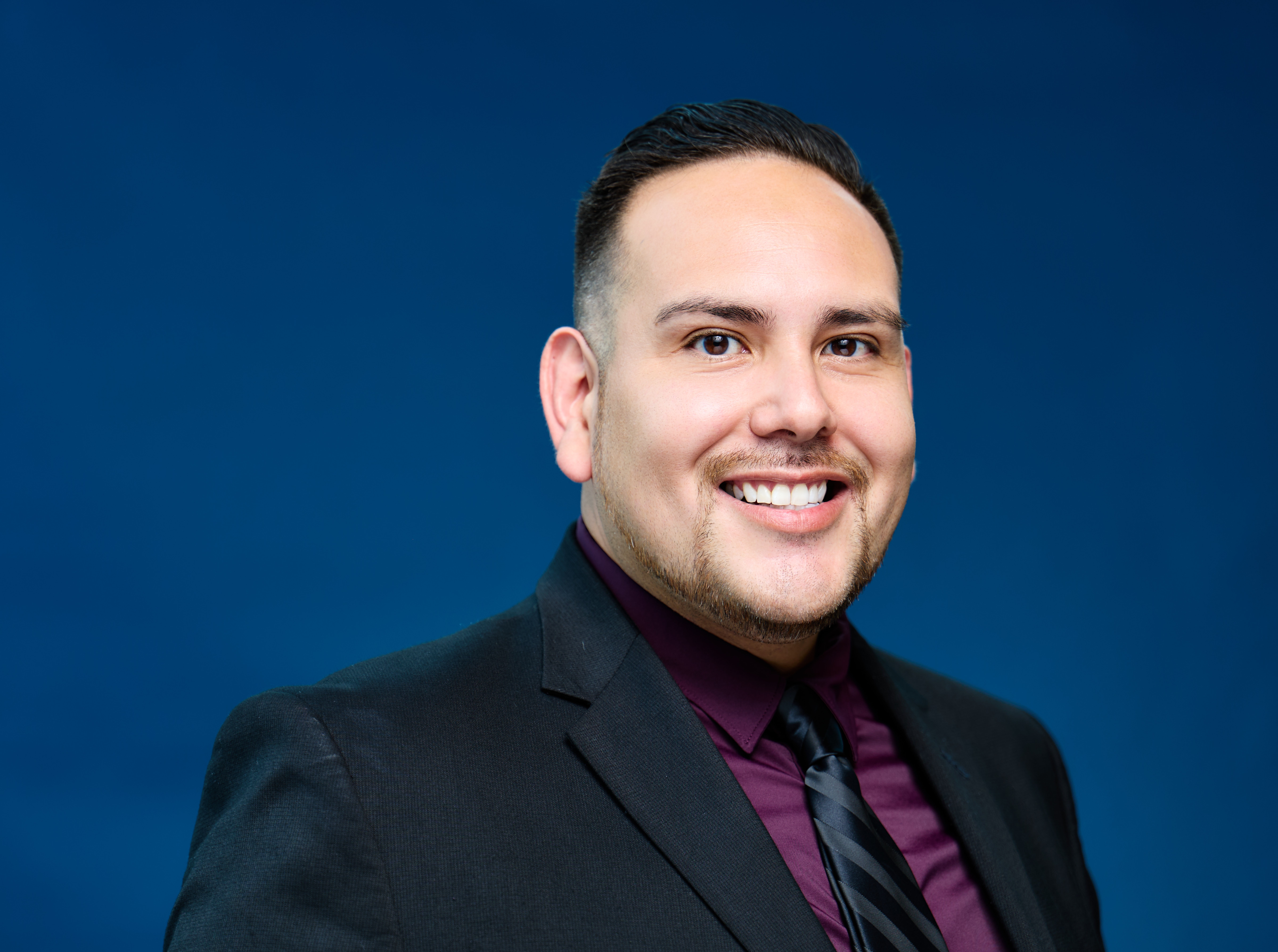 Dr. Manuel Del Real smiling in a black blazer and a dark blue back drop.