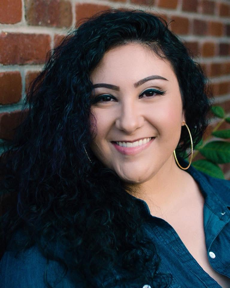 Marianna smiling in a blue shirt in front of a brick wall. 