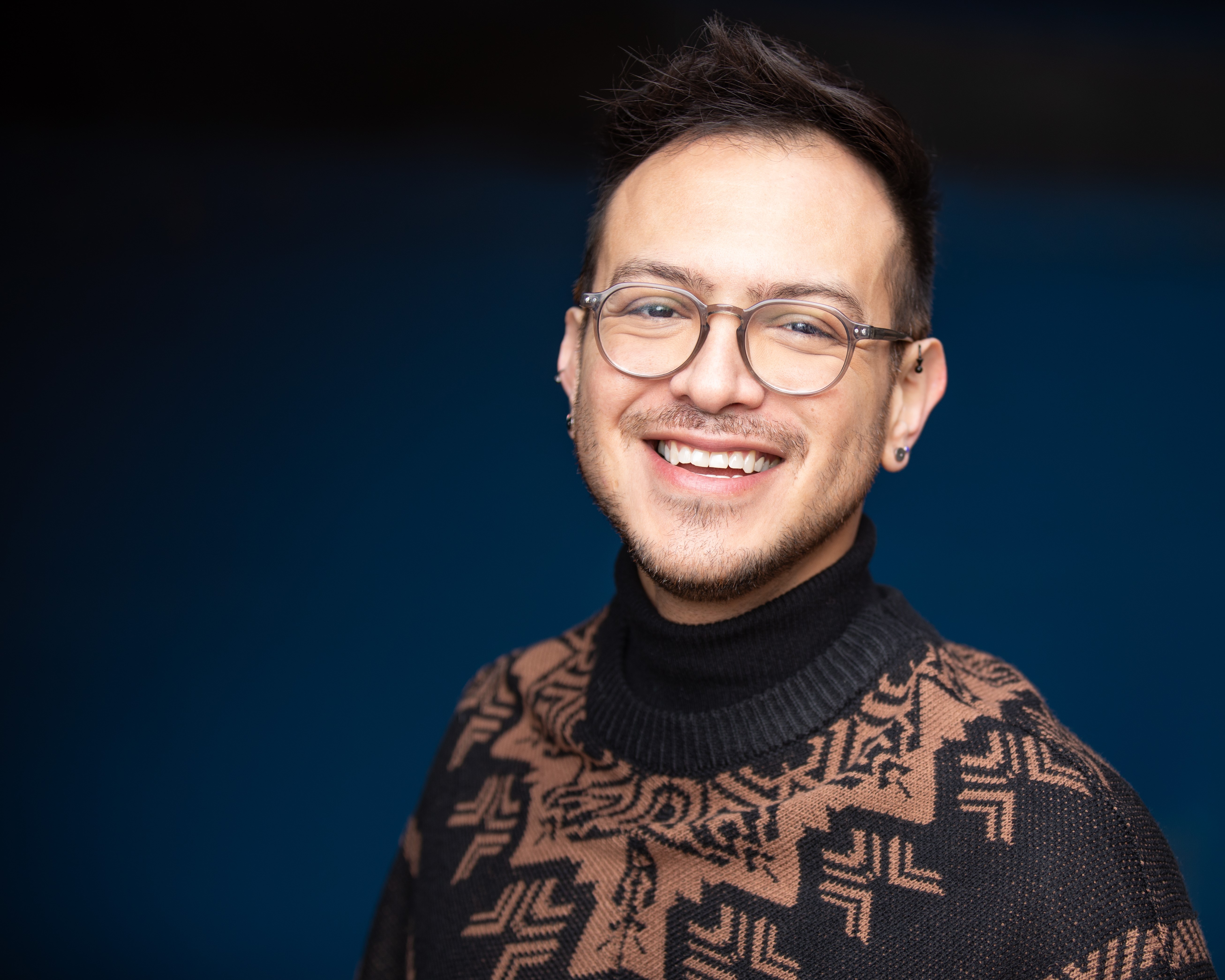 Rick smiling with grey, circle-framed glasses and a pattered sweater against a dark blue backdrop.
