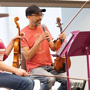 A person seated with a violin speaks into a microphone during the Henry Hoke residency discussion, while other musicians and instruments are partially visible nearby.