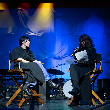 Two people sit on stage in director-style chairs during a live conversation event. One person speaks into a microphone while reading from papers, and the other smiles while listening. Blue stage lighting and musical instruments are visible in the background.