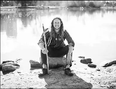 black and white image of Jonathon Stalls, a man with long wavy hair in a striped shirt and dark pants, sitting in front of a still body of water.