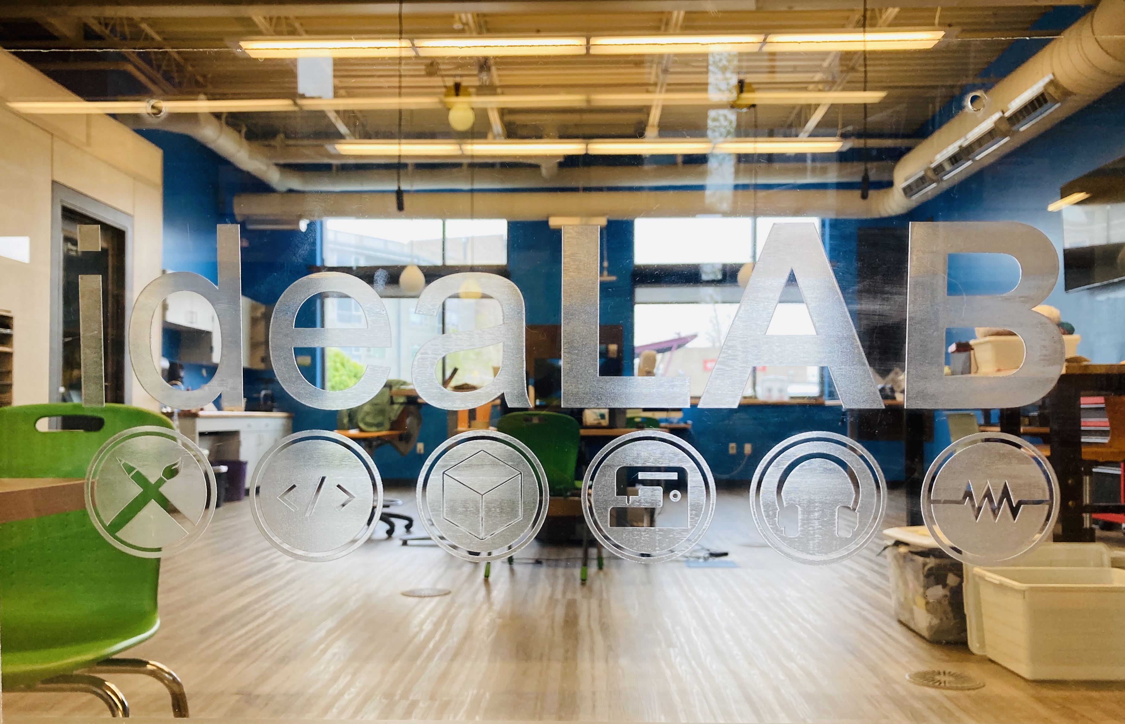 Close up of the word "ideaLAB" etched on to transparent acrylic. Through it, one can see the tables, chairs, and computers inside the Sam Gary Branch ideaLAB.