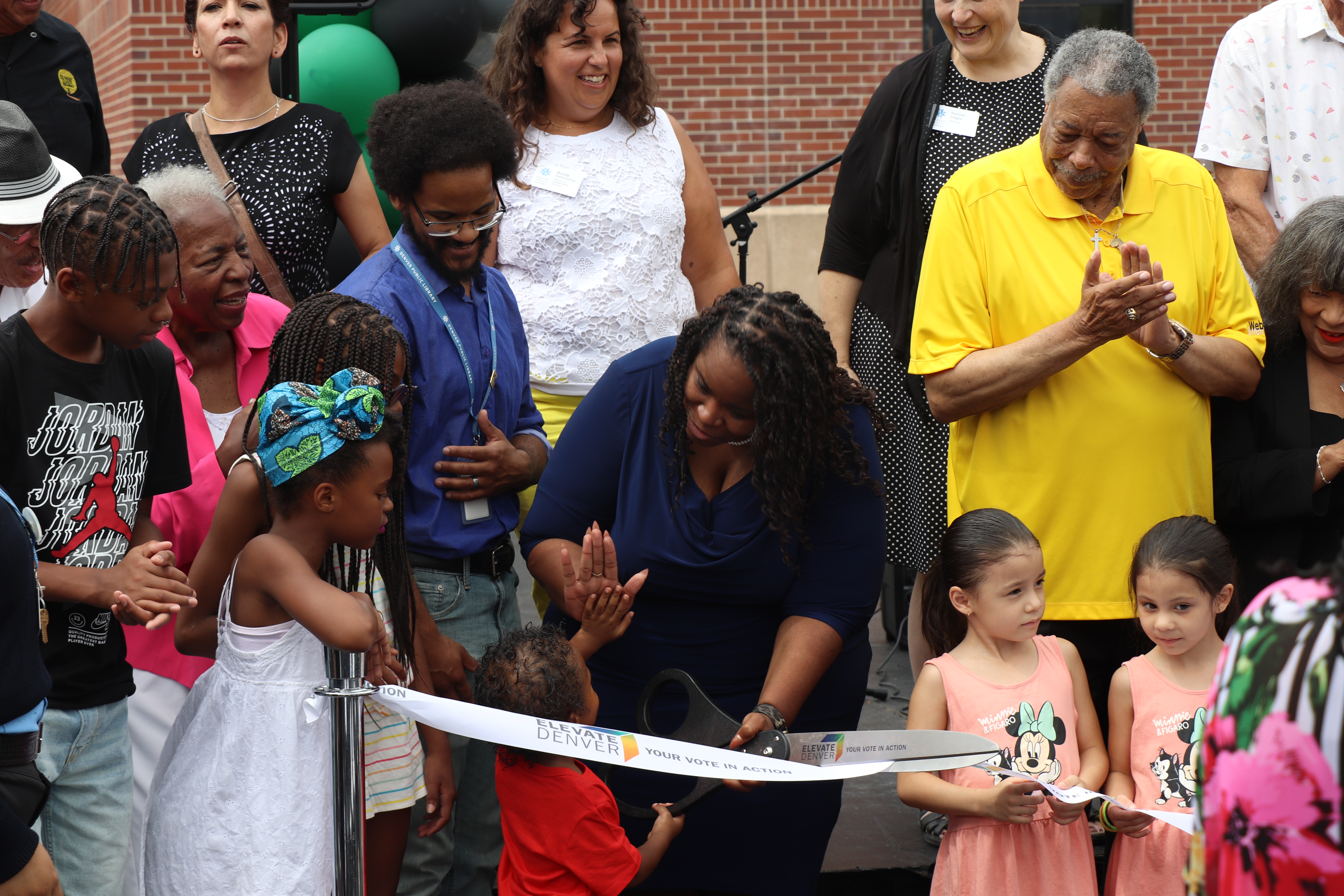 Library staff, dignitaries, and library goers celebrate the reopening of the Blair-Caldwell library with a ribbon cutting ceremony. People are gathered behind a ribbon outside the library and are smiling, high fivng, and cheering.