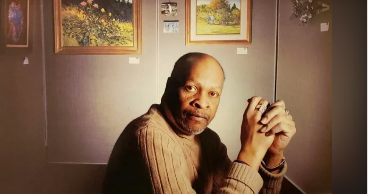 Portrait of Bob Ragland sitting among his artwork hanging on a wall. 