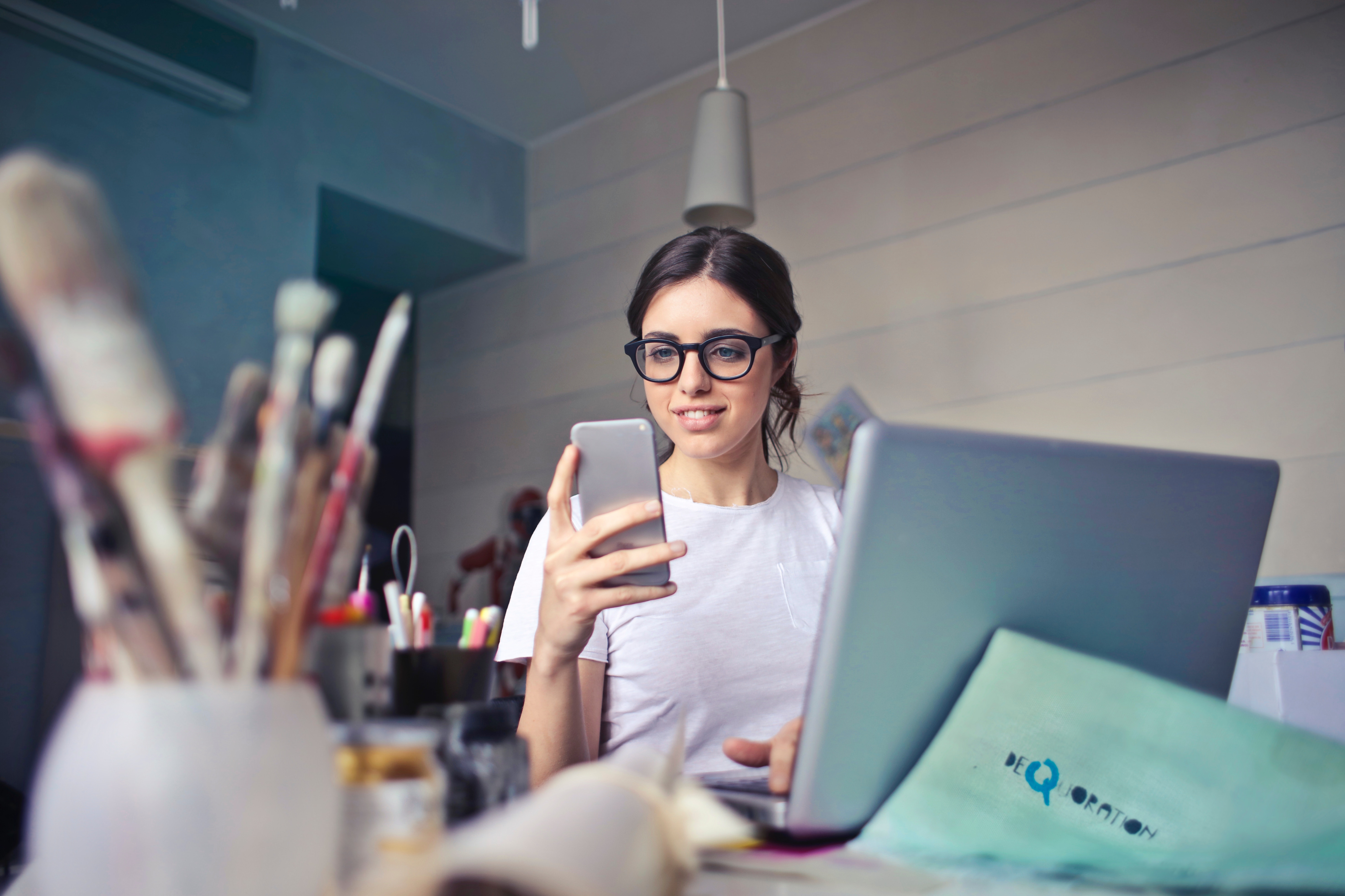 woman in white shirt at desk with computer and paintbrushes using smartphone