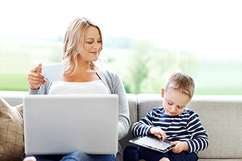 A woman with a laptop and a young boy with a tablet sit together on a sofa