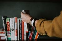 An arm in a yellow sweater with bracelets selects a book from a bookshelf