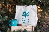 A canvas tote bag with the Winter of Reading logo and a blue mug with the Winter of Reading logo on a box in the woods.
