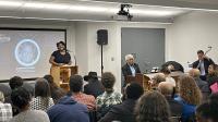 Photograph of one of the award ceremony hosts speaking to room filled audince at Ford-Warren branch. 