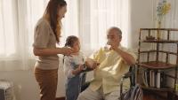 A photo of an older father in a wheelchair, his adult daughter, and granddaughter; he is taking medication with water..