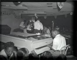 A black and white photo of the Rainbow Ballroom stage. There is a Black man playing piano and a band guddled together holding string instruments.