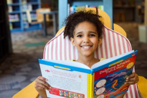 Child smiling at camera, sitting in chair and reading book