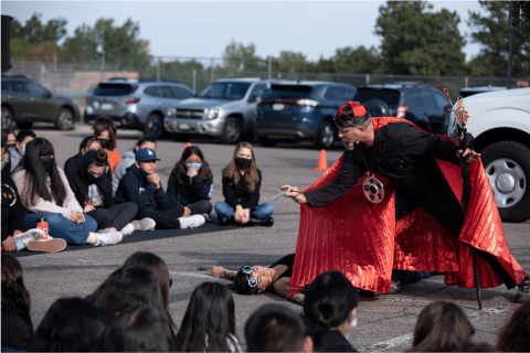 A group of people sit on the ground in a parkinglot surrounded by two actors. One actor is laying on the ground and the other is wearing a red cape and standing above them.