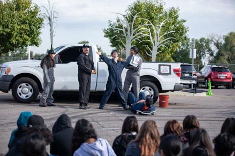 Color photograph of four actors standing in front of a white pickup truck. They are wearing grey and navy blue coveralls. One in the middle has her arms stretched wide and is holding a clipboard.