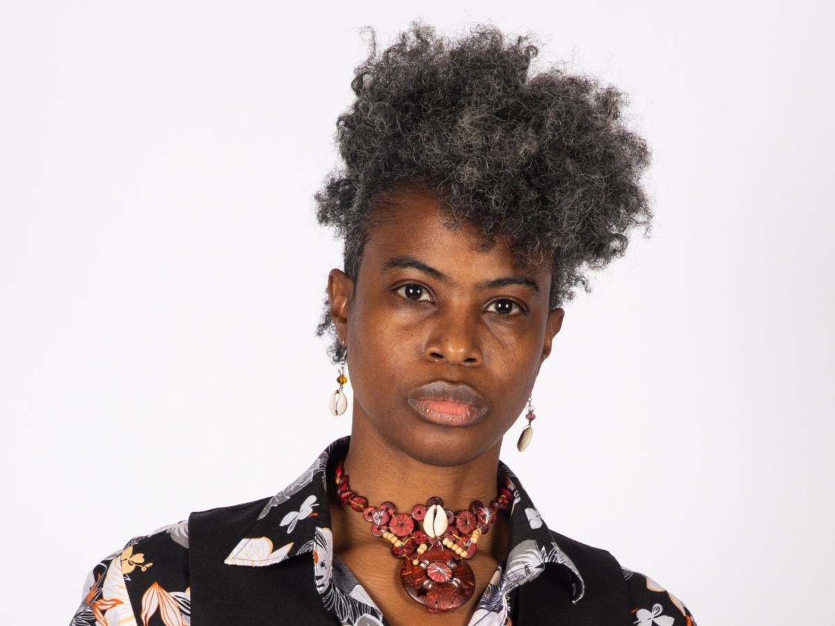 photograph of Adri Norris, a woman with dark skin, short black and grey hair, wearing shell earrings, a red necklace, and black patterned shirt