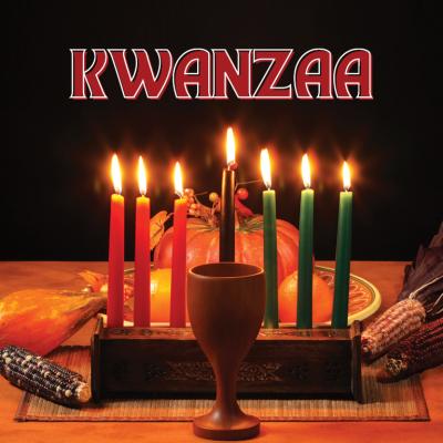 Kwanzaa kinara with seven lit red, green, and black candles and a wooden unity cup on a table decorated with fruits and ears of corn, with the word ‘Kwanzaa’ above.